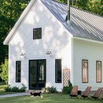 A white, modern farmhouse with a metal roof and black-framed windows sits amidst a green lawn surrounded by trees. Two dogs are playing on the grass near a line of wooden chairs.
