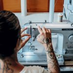 Person with tattoos on their arms adjusting a sewing machine in a workshop.