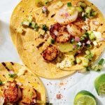 Two corn tortillas topped with grilled scallops, diced cucumber, onion, and salsa, alongside lime wedges and a glass of light beer.