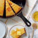 Skillet cornbread cut into wedges, with one piece on a plate topped with butter and honey. Nearby are a honey dipper, jar of honey, and two empty plates on a checkered cloth.