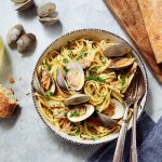 A bowl of clam linguine with parsley garnish, surrounded by a glass of white wine, clams, sliced baguette, and a fork and spoon on a gray surface with a napkin.