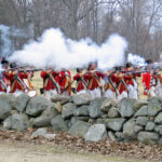 Historical reenactors in red uniforms fire muskets behind a stone wall in an outdoor setting.