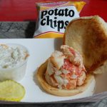 A tray with a lobster roll, a cup of coleslaw, pickle slices, and a bag of potato chips.