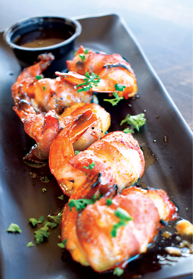 Avanu on Flagler Restaurant, New Smyrna Beach. Three pieces of bacon-wrapped shrimp garnished with parsley are arranged on a black plate with a small cup of dipping sauce.