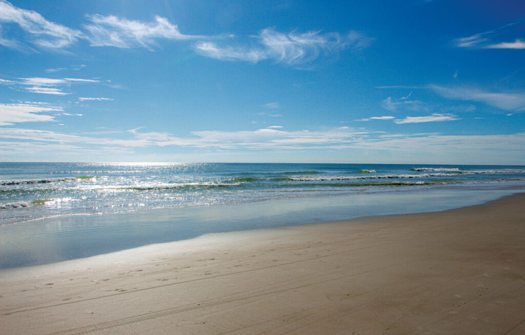 New Smyrna Beach, Florida. A sandy beach with gentle ocean waves under a bright blue sky with scattered clouds. Sunlight reflects off the water, and the shore is mostly empty.