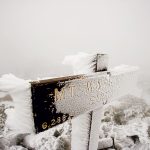 Snow-covered wooden sign at Mt. Washington summit, partially obscured by fog, with rocks and a person in the background.