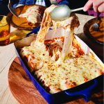 A person lifts a portion of cheesy baked enchiladas out of a blue casserole dish. Nearby are plates and a glass of drink on a wooden table.
