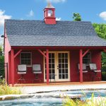 A small red building with a porch, black roof, and cupola stands in front of a fenced-in area with lush greenery and a pond in the foreground. Four chairs are arranged on the porch.