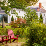 Two red Adirondack chairs are set on a stone patio amid greenery, facing a large white house with multiple windows and a sunroom in the background.
