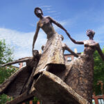 Bronze sculpture of four women holding hands in a circle, outdoors, with trees and brick buildings in the background under a blue sky.