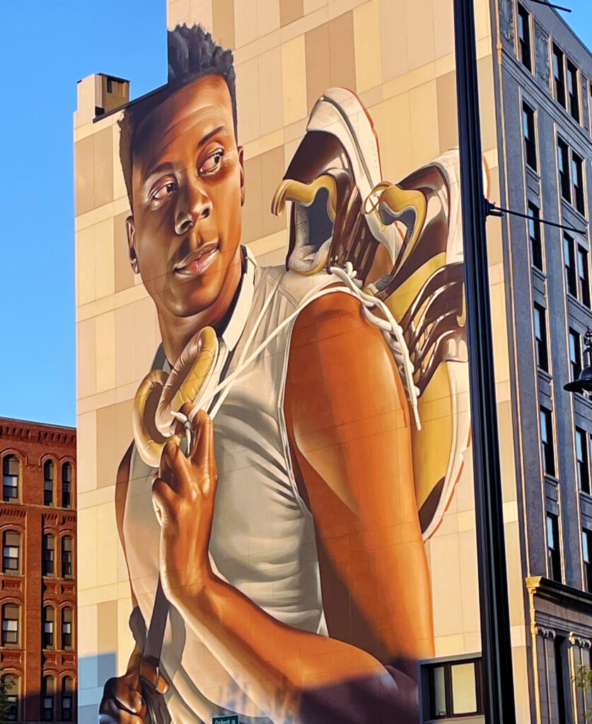 A mural on a building depicts a young man in a white tank top holding a pair of sneakers slung over his shoulder, painted in realistic style.