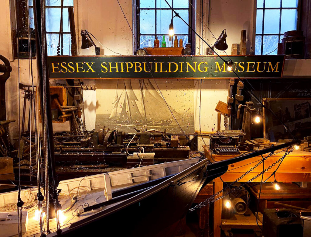 A model ship and antique shipbuilding tools displayed inside the Essex Shipbuilding Museum, with a green and gold sign above.