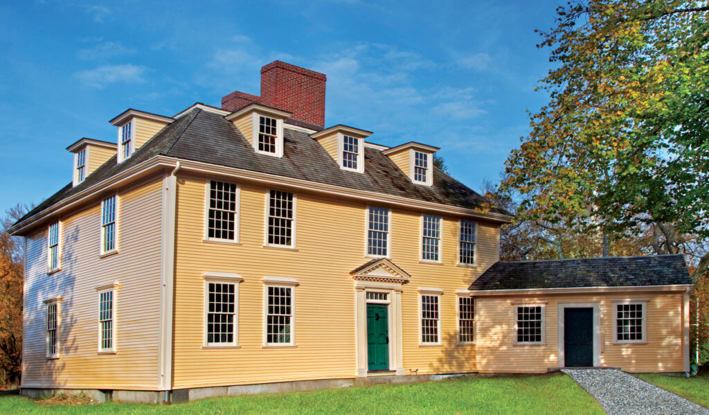 A large, yellow, two-story colonial-style house with multiple dormer windows, a central chimney, and an attached single-story wing, set on a grassy lawn.