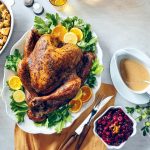 Roast turkey garnished with lemon slices and herbs, surrounded by stuffing, cranberry sauce, and gravy on a table.