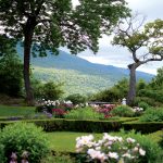 A scenic garden with colorful flowers and neatly trimmed hedges, framed by two large trees. In the background, rolling hills and a cloudy sky are visible.