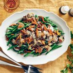 A white platter of green beans topped with sautéed mushrooms and crispy onions on a yellow tablecloth, with cutlery and a small green plant beside it.