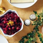 A bowl of cranberry sauce garnished with orange zest is on a table with a gold spoon, salt and pepper shakers, a green leaf branch, and a glass with ice on a mustard-colored tablecloth.