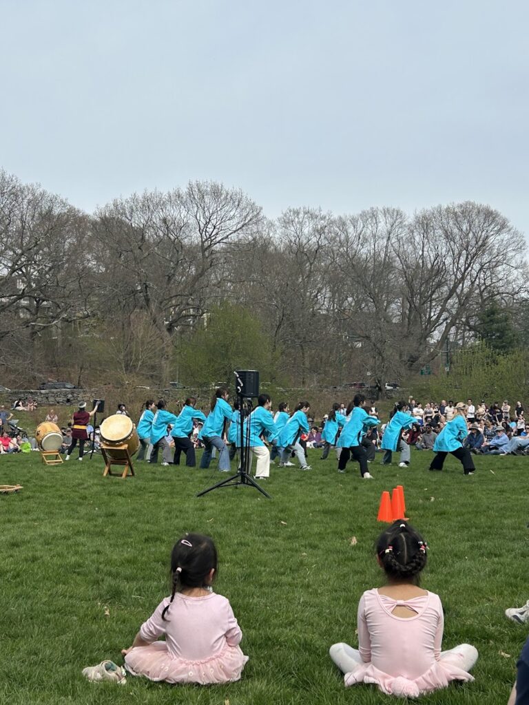 Children in pink outfits sit on grass watching a group in blue jackets perform with drums outdoors; large audience and bare trees in the background.