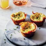 Three egg muffins garnished with chives sit on a round marble platter. A glass of orange juice, a folded napkin, and a glass with a pink beverage are in the background on a marble surface.
