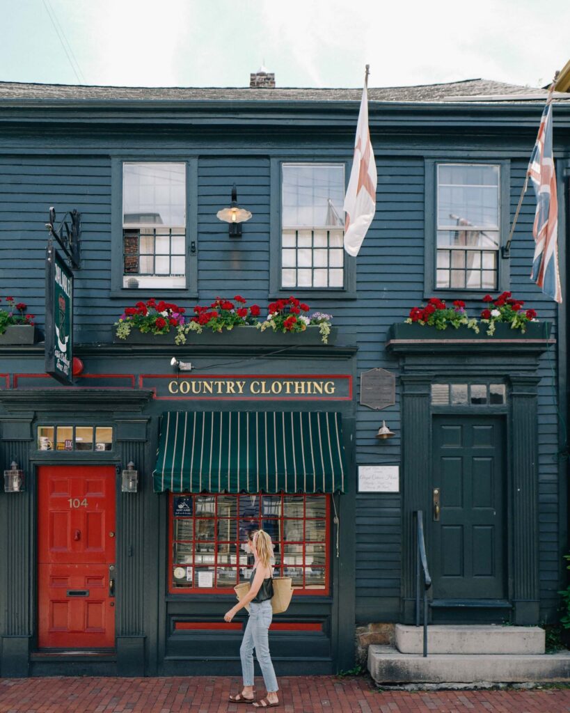 A woman walks past a blue two-story building with flower boxes, striped awning, and a sign that reads "Country Clothing." Flags are displayed on either side of the entrance.
