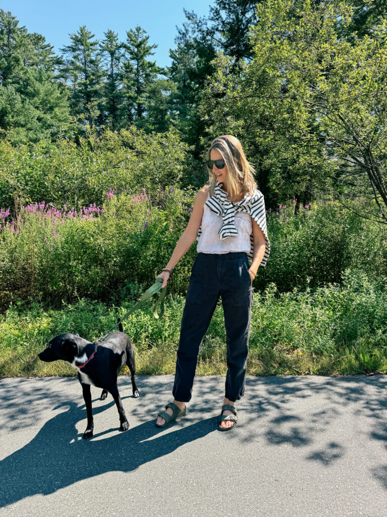 A woman wearing sunglasses and casual clothing stands on a paved path holding a green leash attached to a black dog, with trees and greenery in the background.