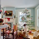 Living room with Christmas tree, two dogs, and a fireplace. Decor includes red accents, a chandelier, and a rug. Natural light fills the room.