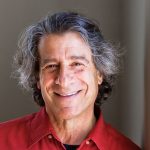 A smiling man with gray hair, wearing a red shirt, stands against a light-colored wall.