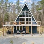 A-frame house with large front windows, elevated deck with seating, outdoor stairs, and surrounded by trees in a wooded area.