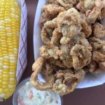A serving of fried seafood in a paper tray, accompanied by a cob of corn and a portion of coleslaw in a small container.