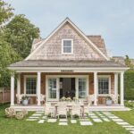 Coastal Charm Behind the Hedge: A Featured Cottage from "Nantucket Looms: A Legacy of Style" - Cozy house with wooden shingles, columns, and a front porch. A dining table with chairs is set on the lawn, surrounded by hedges and a tree.