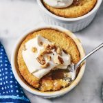 A ramekin of peach cobbler topped with whipped cream and chopped walnuts, with a spoon taking a portion. Two other ramekins and three spoons on a blue dotted napkin are nearby.