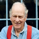 Elderly man smiling, wearing a blue shirt and red suspenders, standing in front of a window.