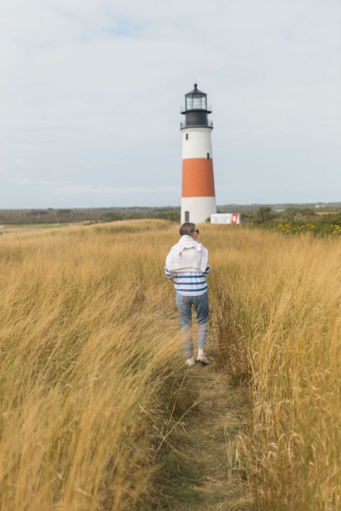 A person walks along a grassy path toward a tall, white and red-striped lighthouse under a cloudy sky.