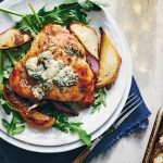 Plate of roasted chicken topped with blue cheese crumbles, served on a bed of arugula with roasted pears and onions, next to a small tray of roasted vegetables.