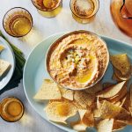A bowl of hummus with olive oil garnish on a plate with tortilla chips. Accompanied by a stack of napkins, an empty plate, several glasses, and a pitcher on a white surface.