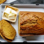 A loaf of pumpkin bread with two slices cut, served on a white tray next to a small dish of butter.