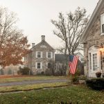 Stone house with an American flag by the front door, trees with autumn leaves, and cloudy sky. Nearby road and another stone building in the background.