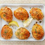 Six stuffed quahogs topped with herbs on a white tray, garnished with two lemon wedges, set on a woven surface.
