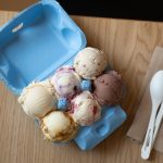 Six scoops of assorted ice cream are arranged in a blue egg carton on a wooden table, next to two white plastic spoons and brown napkins.
