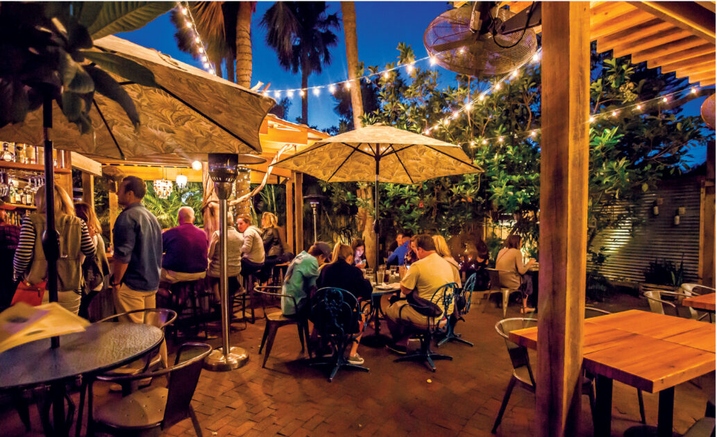 Third Wave restaurant, New Smyrna Beach. People sit at tables and under umbrellas in a warmly lit outdoor restaurant patio at dusk, with string lights overhead and trees in the background.