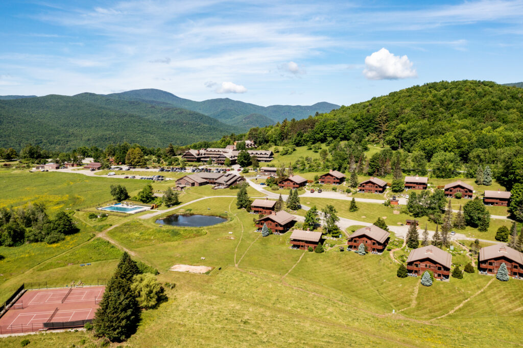 Aerial view of a rural resort with cabins, a swimming pool, pond, tennis court, and wooded mountains in the background under a partly cloudy sky.