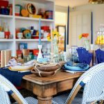 A dining table set with plates, candles, and glassware. Blue and white chairs surround the table. Shelves in the background hold various colorful dishes and items.
