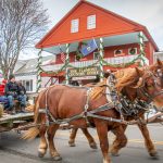 A horse-drawn carriage with people rides past The Vermont Country Store, decorated with flags and garlands, on a snowy day.