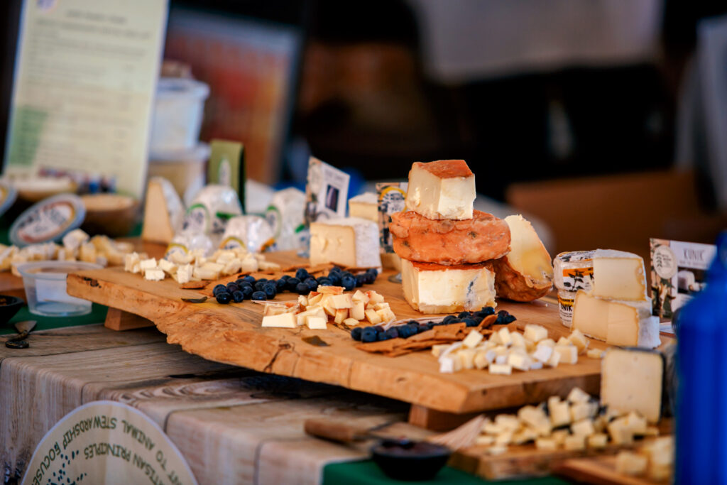 A wooden board displays various types of cheese, cut into cubes and wedges, with scattered blueberries and cheese labels on a table.