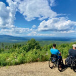 Accessible Travel in New England: Top Inclusive Adventures for a Barrier-Free Summer: Two people in wheelchairs sit on a dirt path overlooking a lush green valley with mountains and a cloudy sky in the background.