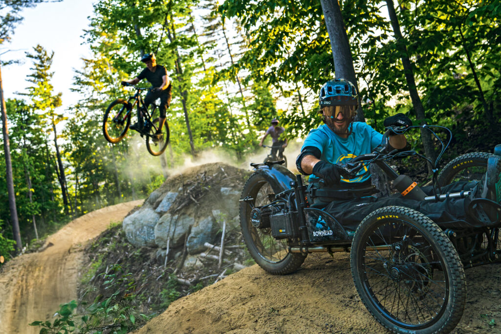 Accessible Travel in New England: Top Inclusive Adventures for a Barrier-Free Summer: Three people ride adaptive and standard mountain bikes on a dirt trail through a forest; one is airborne while another navigates a turn in an off-road wheelchair bike.