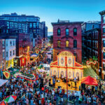 A large crowd gathers at an outdoor street festival with colorful lights, food stalls, and a decorated stage set between brick buildings at dusk.