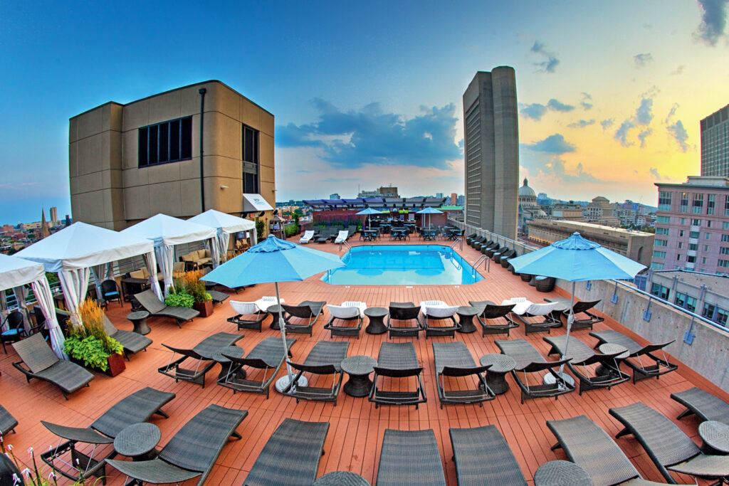 Best Things to Do in Boston This Summer . Rooftop pool area with lounge chairs, umbrellas, and cabanas overlooking a cityscape at sunset.