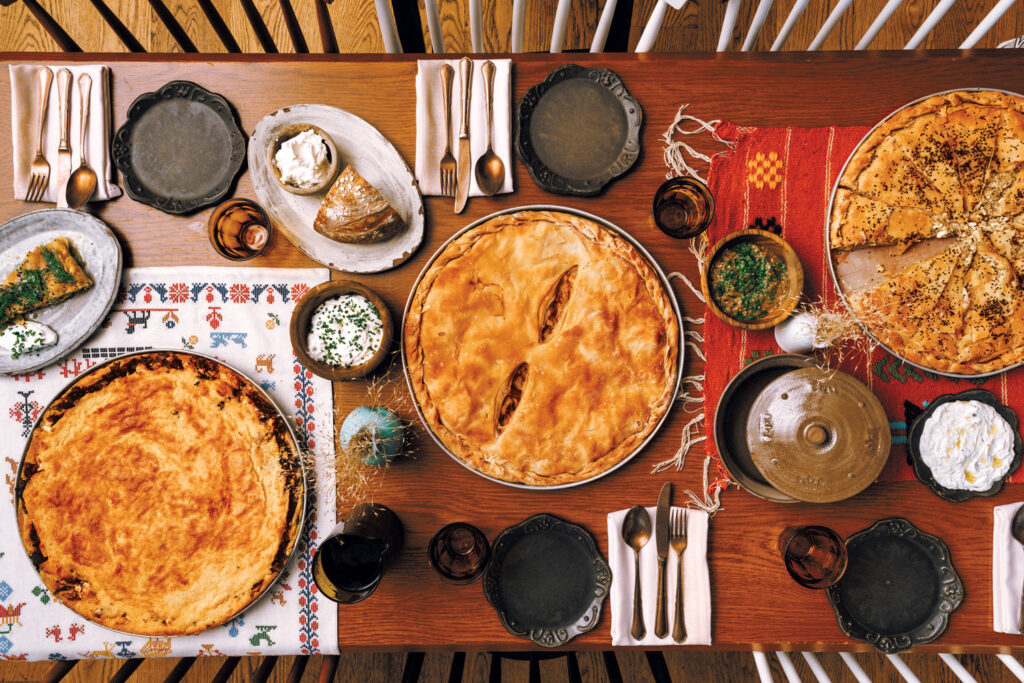 A table set for four with plates, utensils, glasses, various pies, side dishes, bread, and spreads on patterned tablecloths.
