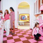 Three people in a retro-style pink kitchen; two stand at the counter, one crouches to check food in the oven. The room has pink appliances, pink and brown checkered floor, and a doorway.